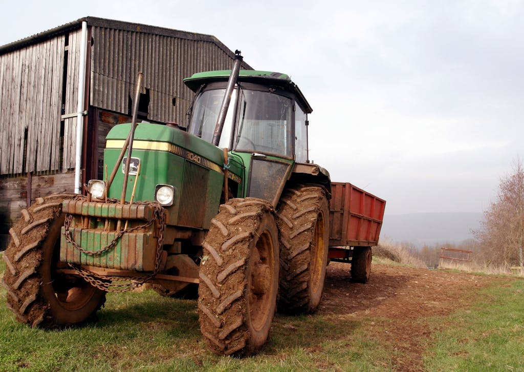 A green tractor with a trailer parked beside a rustic barn in Barrow Gurney, UK, during a cloudy day.