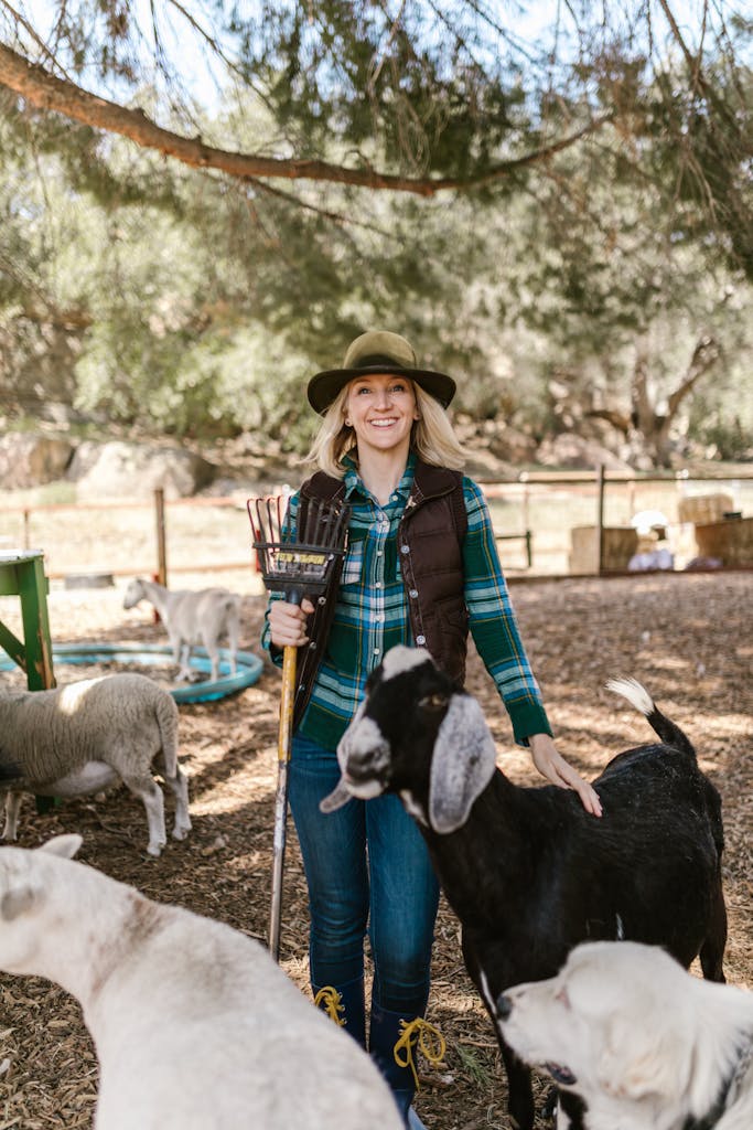 Cheerful woman in cowboy hat standing with goats on a sunny farm day.